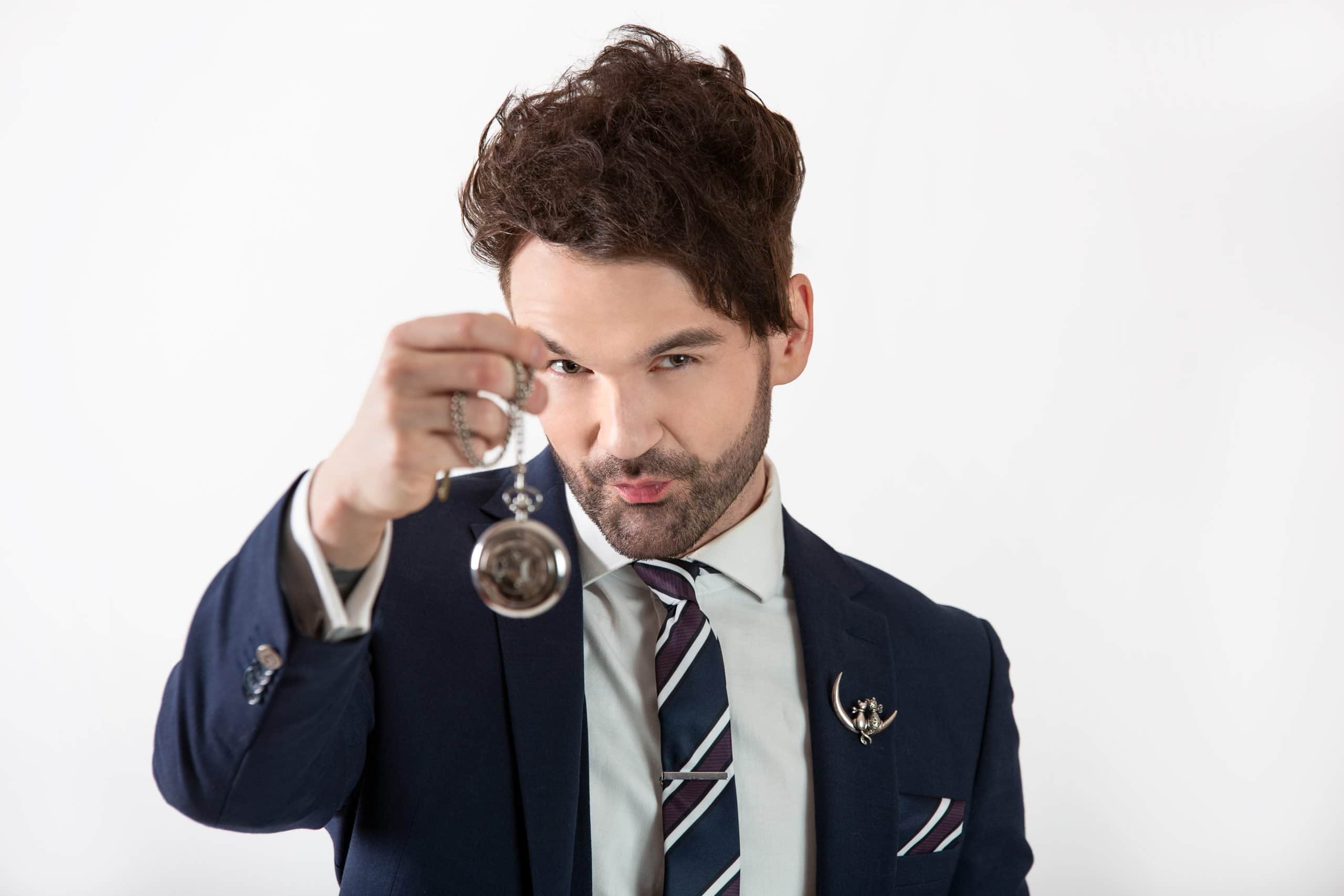Colin Cloud, a mentalist, holding a pocket watch and engaging the viewer with a confident expression.