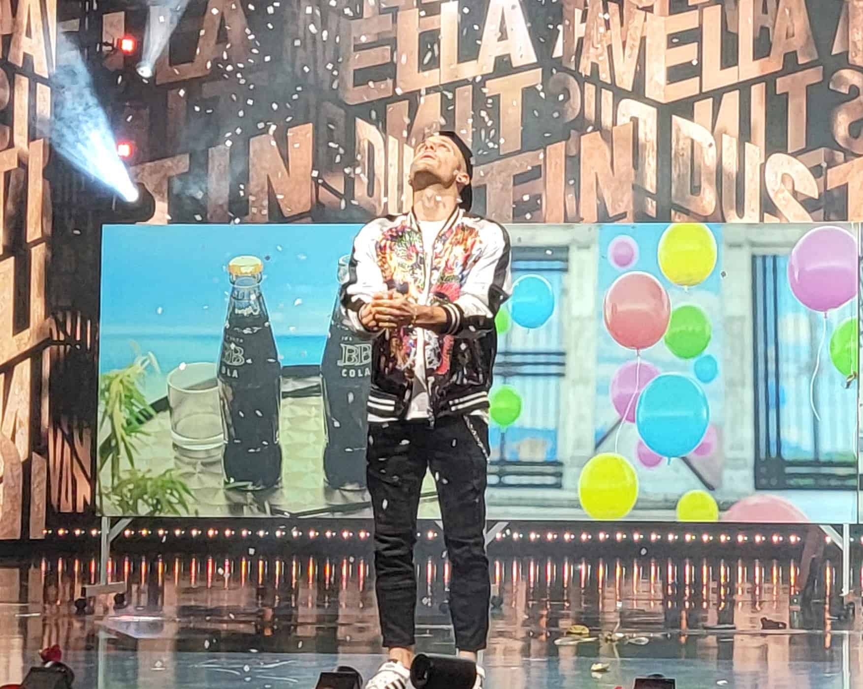 Dustin Tavella performing on stage during America's Got Talent, surrounded by confetti and colorful visuals in the background.